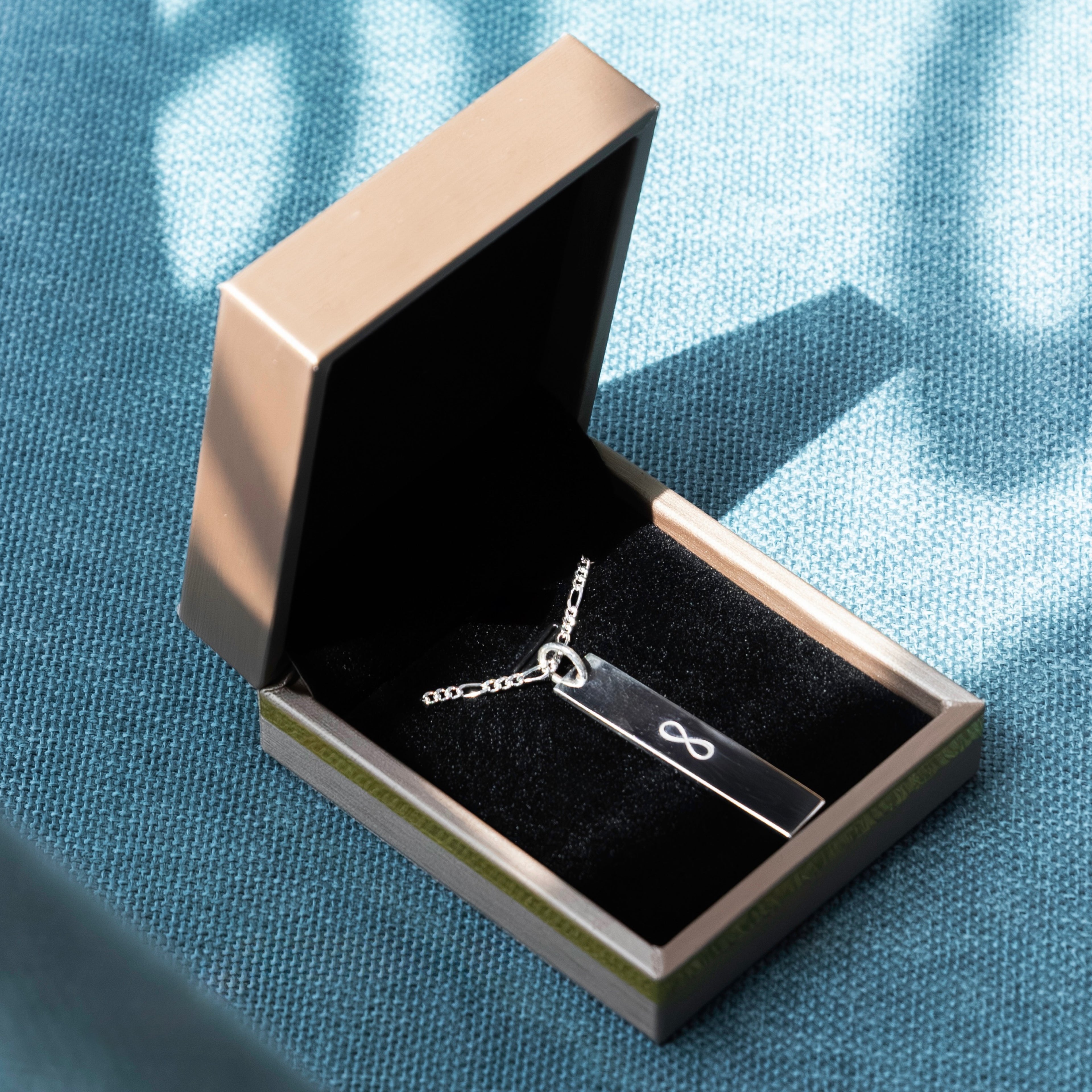 Silver necklace with a dog tag featuring an infinity symbol in an open jewelry box on a blue fabric background.