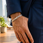 A close-up of a man's hand wearing a silver chain link bracelet, dressed in a navy blue suit jacket and trousers, with a blurred outdoor background featuring a wooden deck and potted plants.