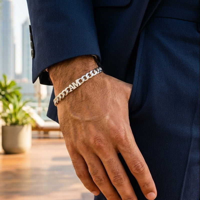 A close-up of a man's hand wearing a silver chain link bracelet, dressed in a navy blue suit jacket and trousers, with a blurred outdoor background featuring a wooden deck and potted plants.